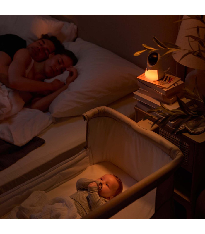 Vigilabebés Kinderkraft Niteeye Glow con la luz cálida encendida en un dormitorio a oscuras junto a la minicuna